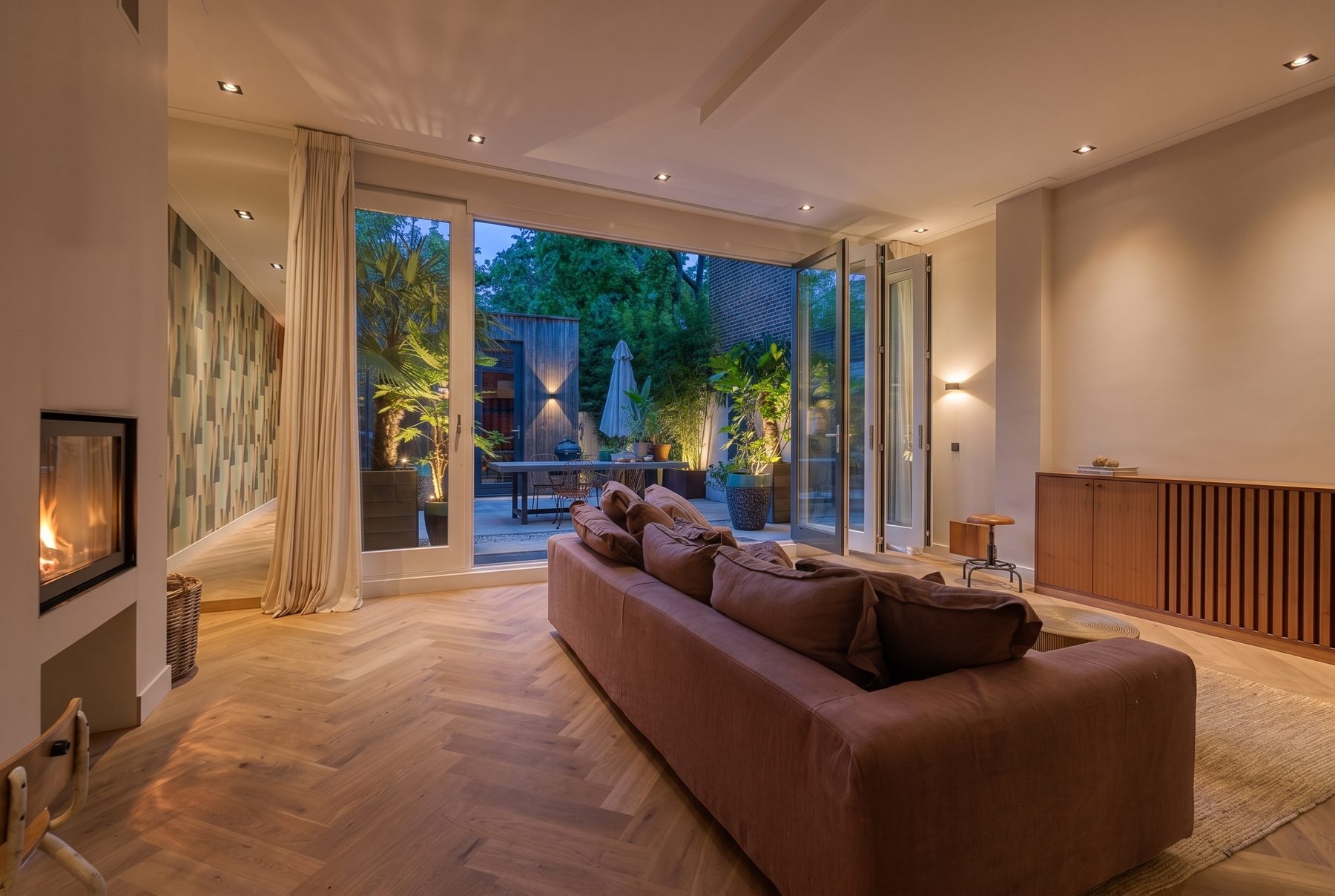 A living room with a fireplace and doors open to the garden at dusk